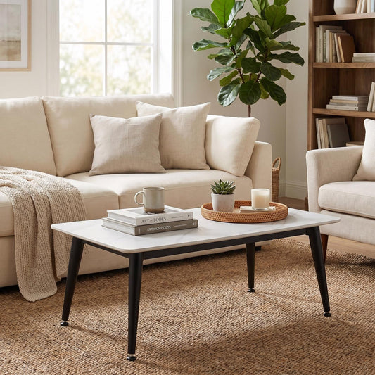 A rectangular coffee table with a white top and black angled legs, styled in a bright living room with a cream sofa, throw blanket, books, and a potted plant.