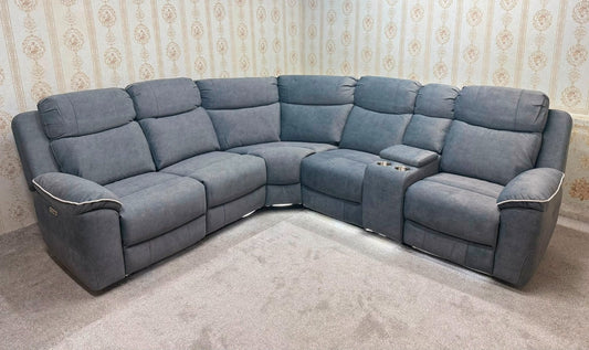 A large L-shaped modular recliner sofa in a dusty blue-gray fabric, positioned in a room with patterned wallpaper and a light-colored carpet.