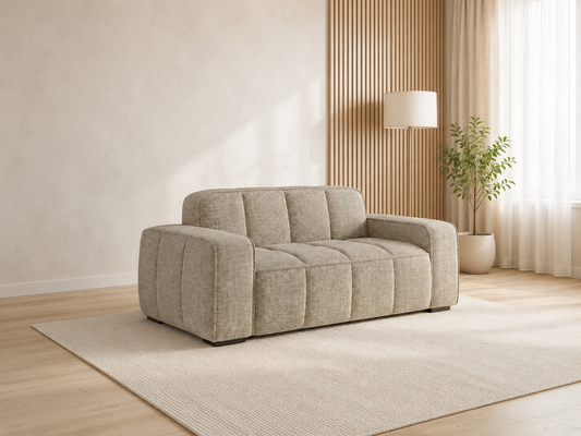 A modern beige upholstered sofa with a chunky, blocky design and tufted back cushions, shown in a minimalist, warm-toned living room setting with a light wood floor, textured rug, wall slat panel, and a potted plant.