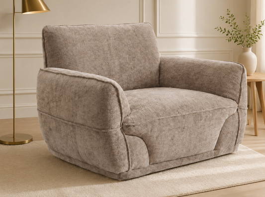 A plush upholstered armchair in a warm taupe-beige textured fabric, styled in a cozy living room setting with a gold floor lamp, a wooden side table, and a beige rug.