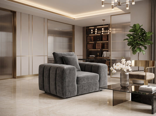 A modern gray velvet club chair with a unique channel-tufted backrest, styled in a luxurious contemporary living room with beige walls, gold trim, and a glass coffee table.