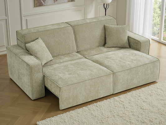 A light sage green upholstered sofa with two matching throw pillows, shown in a bright living room setting with a wooden floor and white rug.