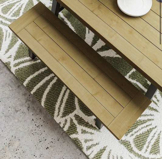 A light natural wood outdoor dining table with a matching bench, shown from an elevated angle on a green and white patterned rug over a concrete floor.