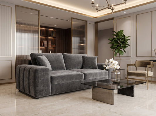 A modern gray velvet sectional sofa in a luxurious, contemporary living room with beige walls, gold trim, a glass coffee table, and a potted plant.