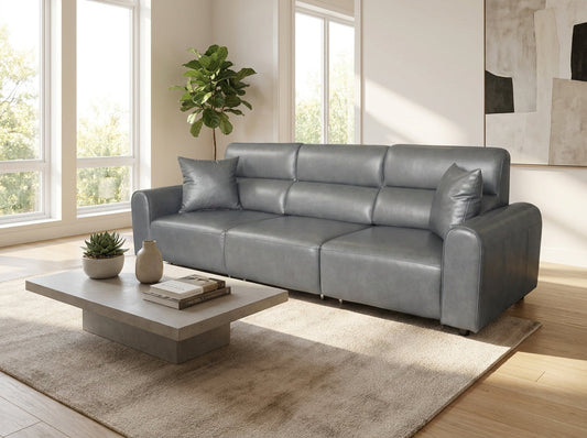 A modern three-seater sofa in a cool gray faux leather, styled in a bright living room with a matching gray throw pillow, a concrete coffee table, and a beige rug.