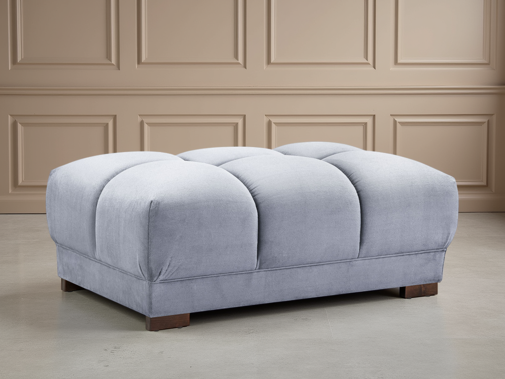 Gray upholstered ottoman on a wooden floor with beige paneled walls.