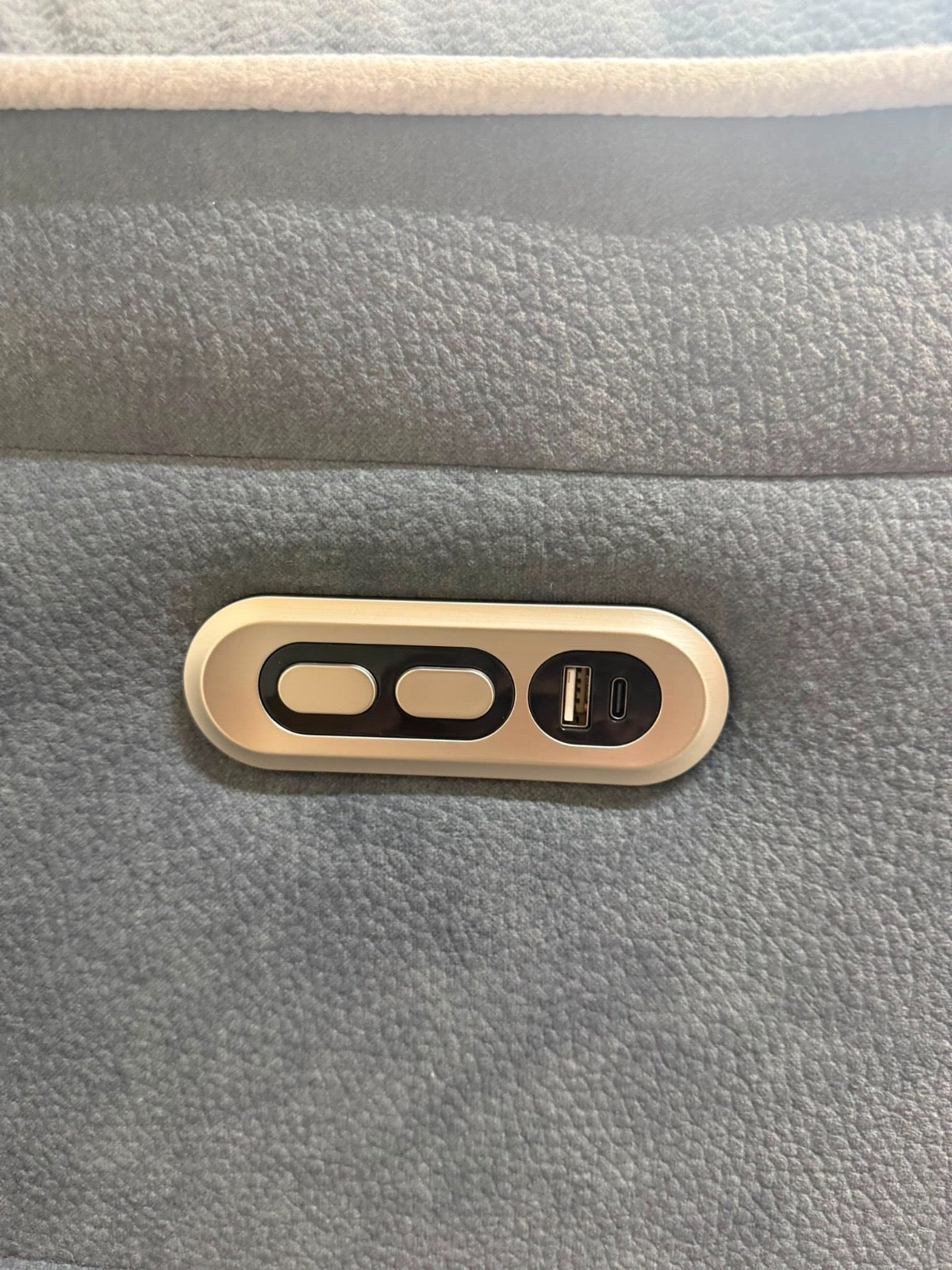 Close-up of a light beige oval control panel with two buttons and a USB-C port, mounted on a textured gray fabric surface.
