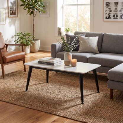 A modern rectangular coffee table with a white top and black angled legs, styled in a bright living room next to a gray sofa and a brown leather armchair.