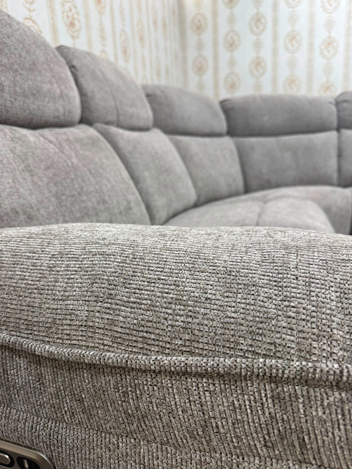 Close-up of a textured gray fabric sectional sofa cushion with tufted channel detailing, showing the woven upholstery in a home setting with patterned wallpaper in the background.