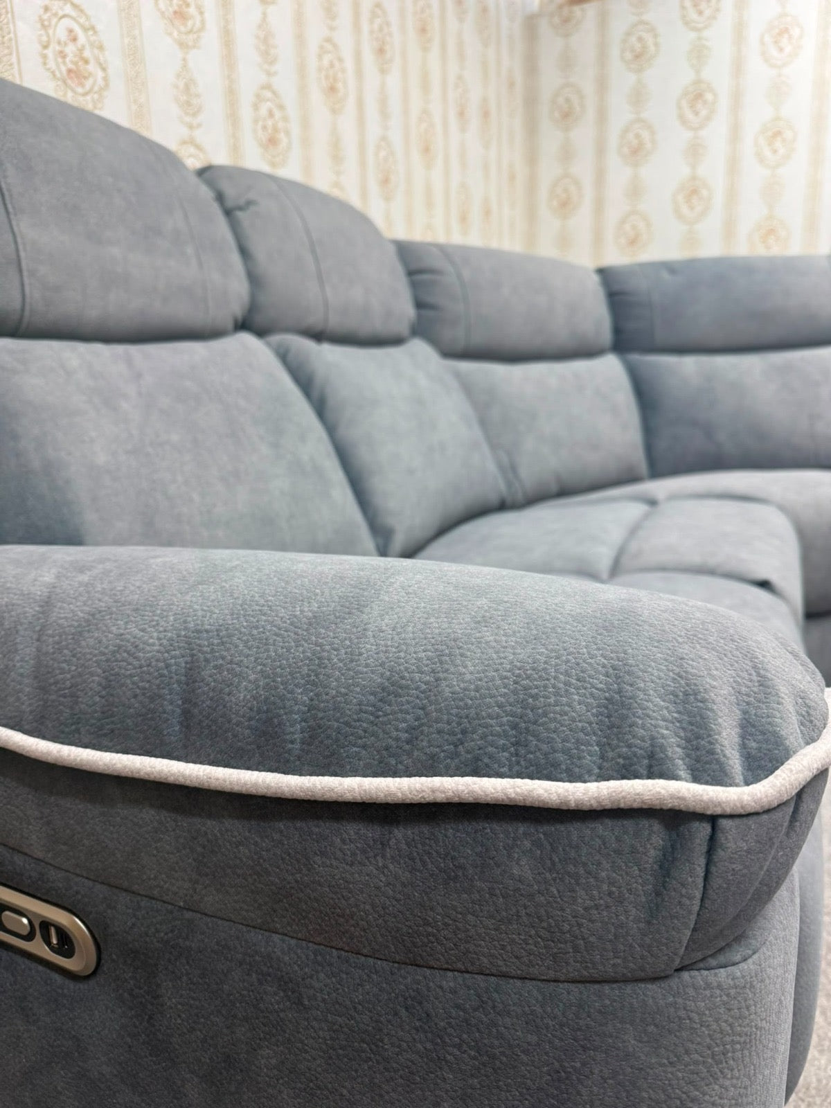 A close-up of a gray fabric reclining sofa with a light-colored piping trim along the armrest, set against a background of patterned wallpaper.
