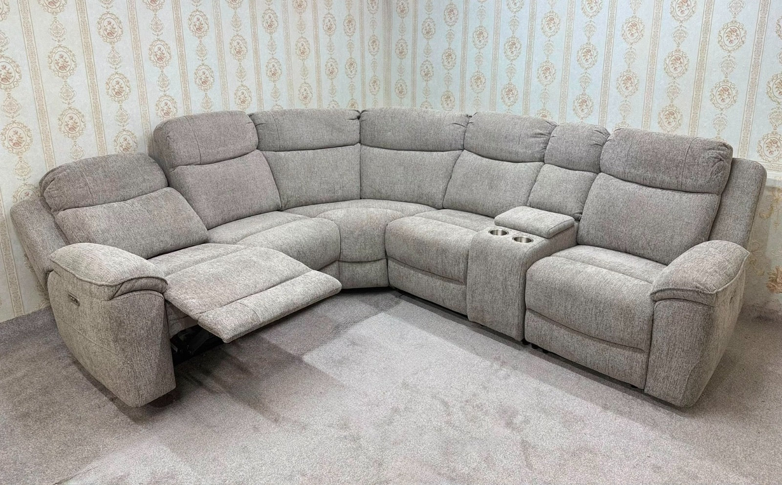 A light gray upholstered sectional sofa with a recliner section on the left with the footrest extended, a center console with cup holders, and a right-facing recliner seat, shown in a room with patterned wallpaper and a gray carpet.