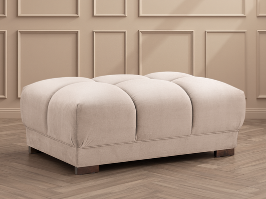 Beige ottoman with tufted design on a wooden floor with panelled walls.