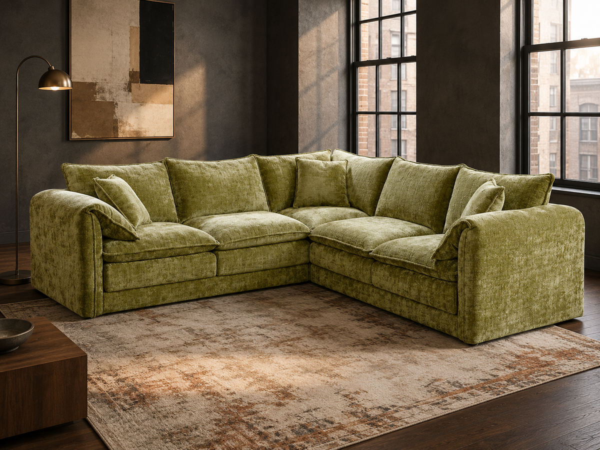 An L-shaped sectional sofa upholstered in textured olive green fabric, styled in a modern loft room with dark gray walls, large black-framed windows, a distressed beige rug, and a wooden coffee table.