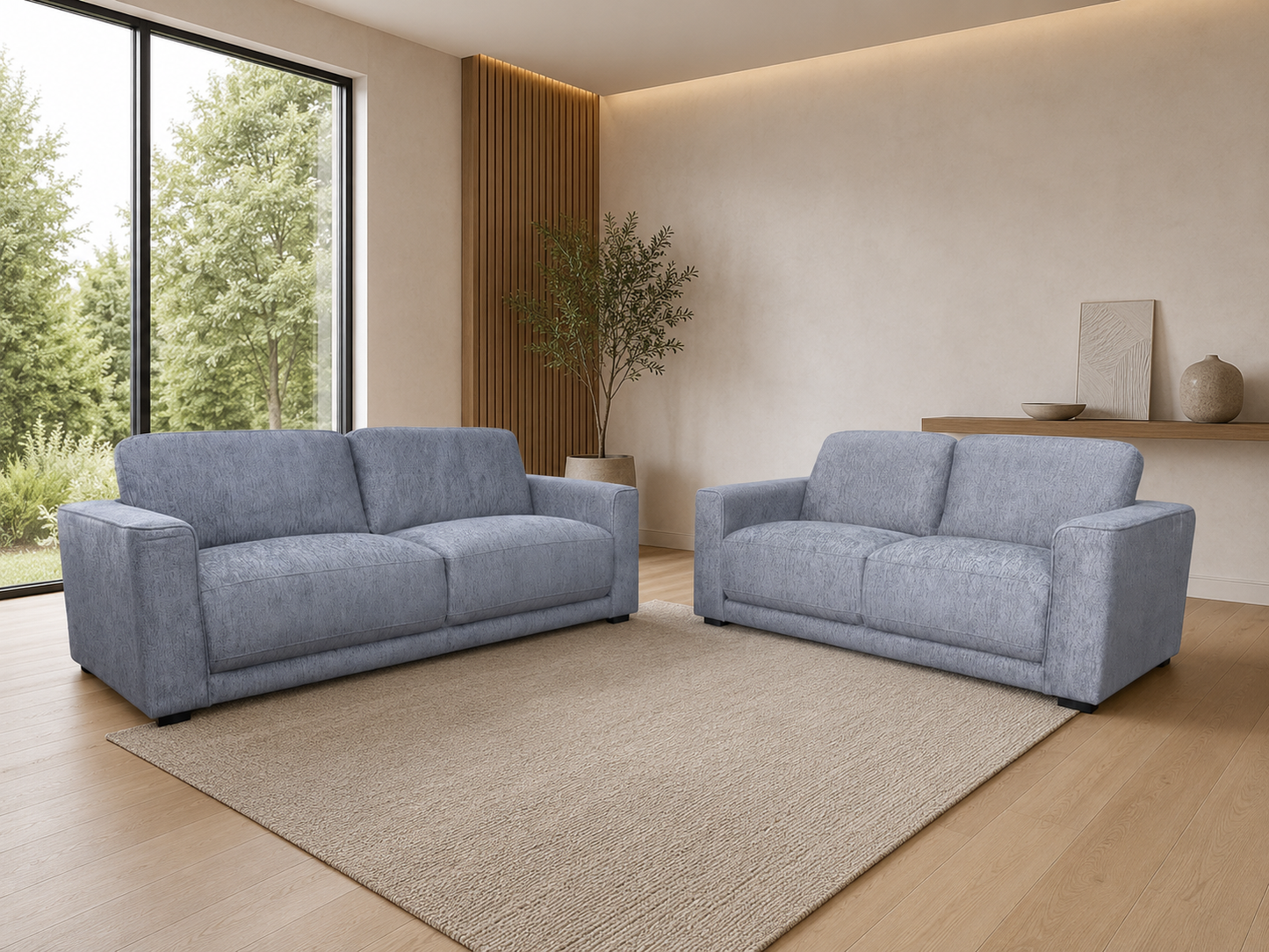 A modern living room setup featuring two light blue upholstered sofas, one larger three-seater and one smaller two-seater, placed on a large beige area rug over light wood flooring. The room has warm beige walls, a vertical wood slat accent, a potted tree, and a floating shelf with decorative items.