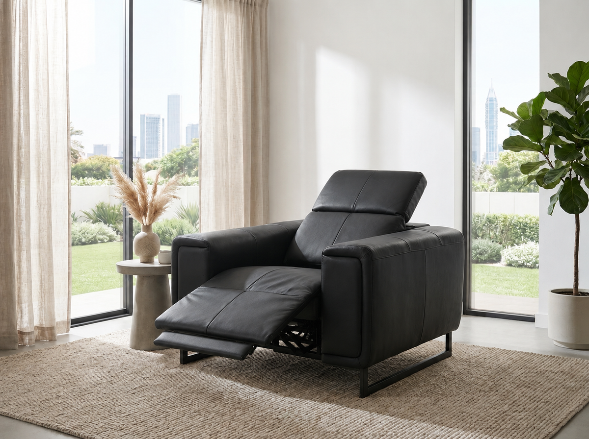 A black leather recliner chair with the footrest extended, positioned on a beige rug in a bright, modern living room with large windows and city views.