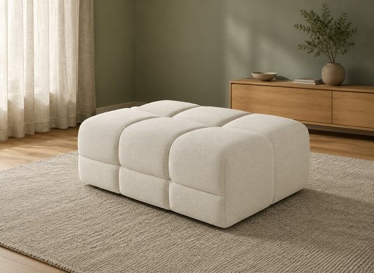 A modern, low-profile ottoman with a tufted grid pattern in a warm off-white fabric, styled in a minimalist room setting on a textured rug.