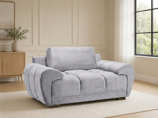 A light gray plush velvet armchair with a tufted seat cushion and rounded arms, shown in a bright, modern living room setting.