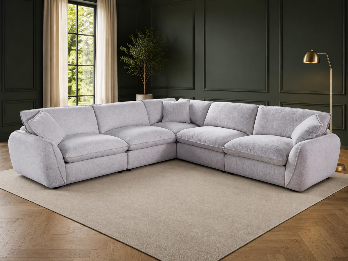A light Beige upholstered sectional sofa arranged in an L-shape on a beige area rug inside a modern room with dark green paneled walls, wood flooring, a window, and a floor lamp.