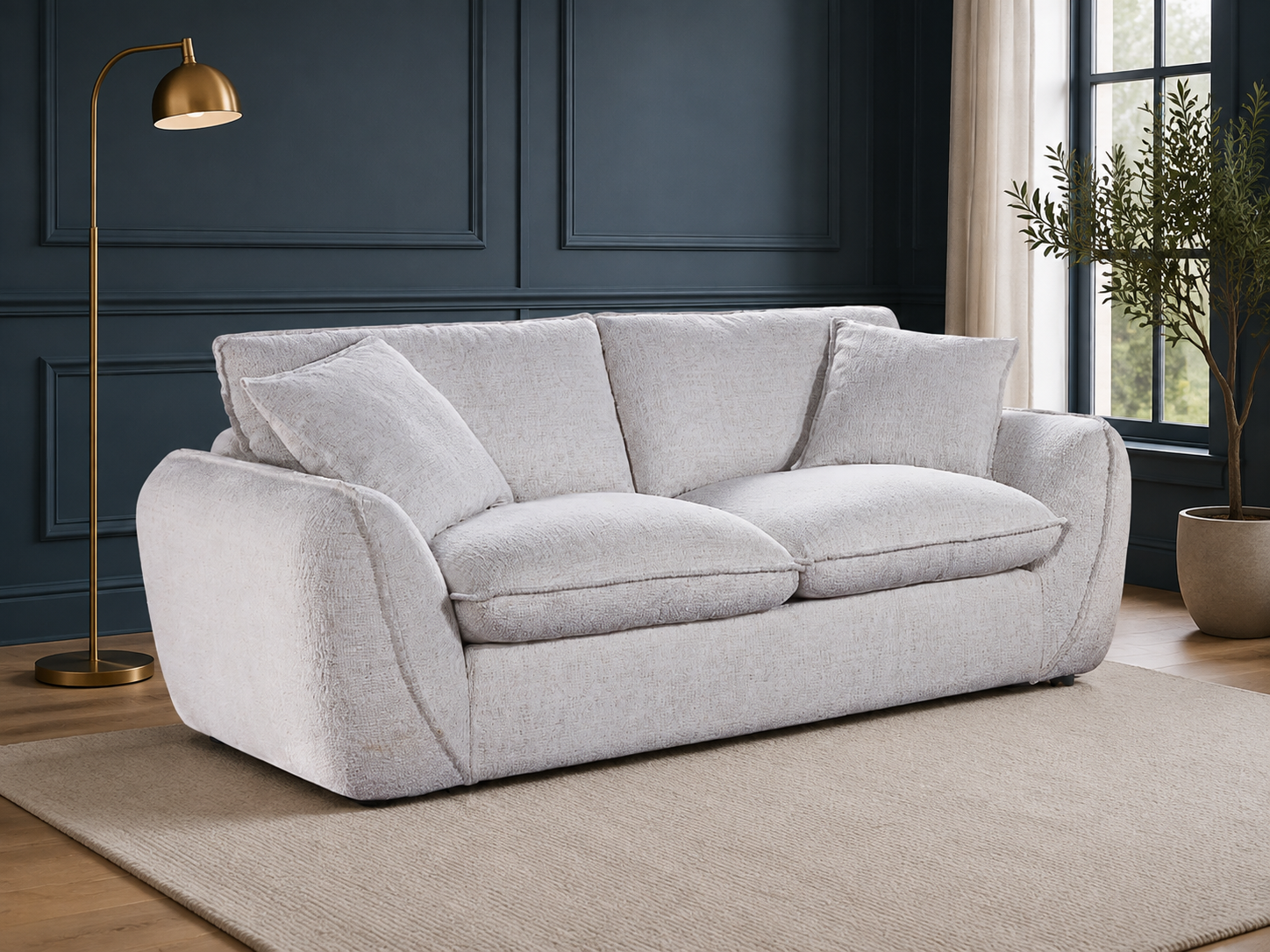 A light beige upholstered loveseat with two loose back cushions and two seat cushions, shown in a styled room with a dark blue paneled wall, a gold floor lamp, a beige rug, and a potted plant.