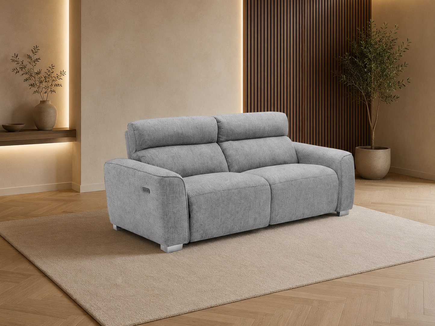 A light gray upholstered reclining loveseat sofa placed on a beige area rug in a modern living room with wood flooring, warm wall lighting, and a potted plant.