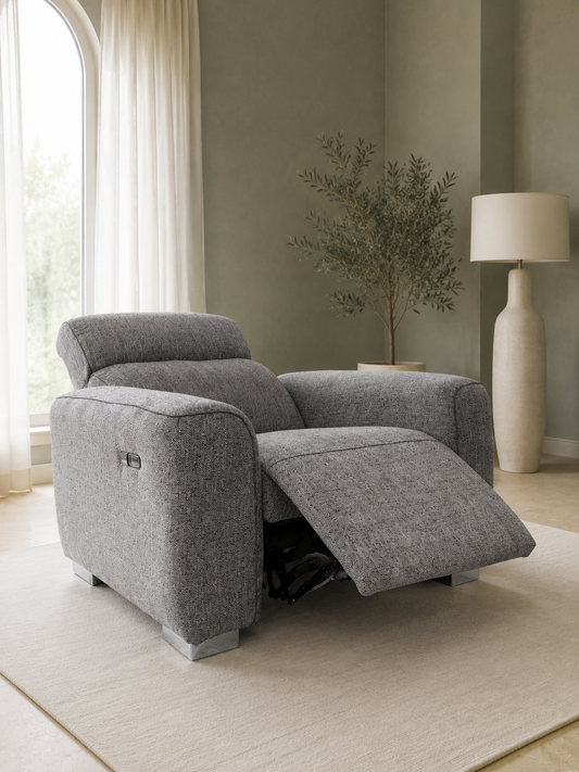 A gray upholstered recliner chair with the footrest extended, shown in a softly lit living room with a rug, floor lamp, curtains, and a potted tree.