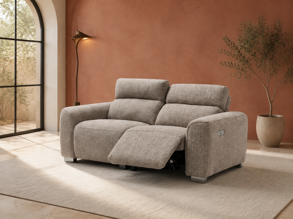 A light gray upholstered reclining loveseat sofa shown in a styled living room with a terracotta wall, floor lamp, arched window, rug, and potted plant.