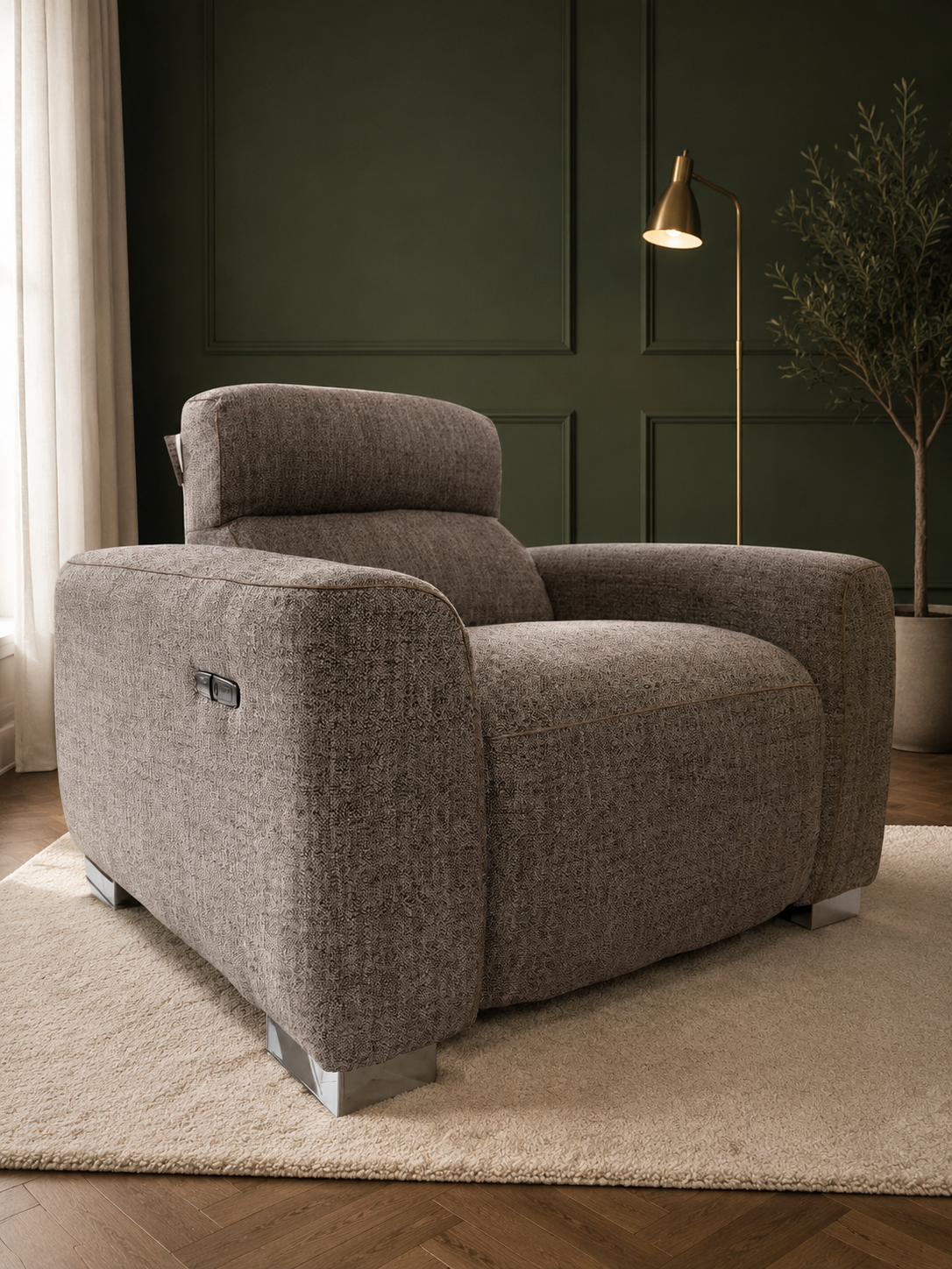 A modern upholstered armchair in textured taupe-gray fabric, positioned on a cream shag rug in a styled living room with a dark green paneled wall, floor lamp, and potted tree.