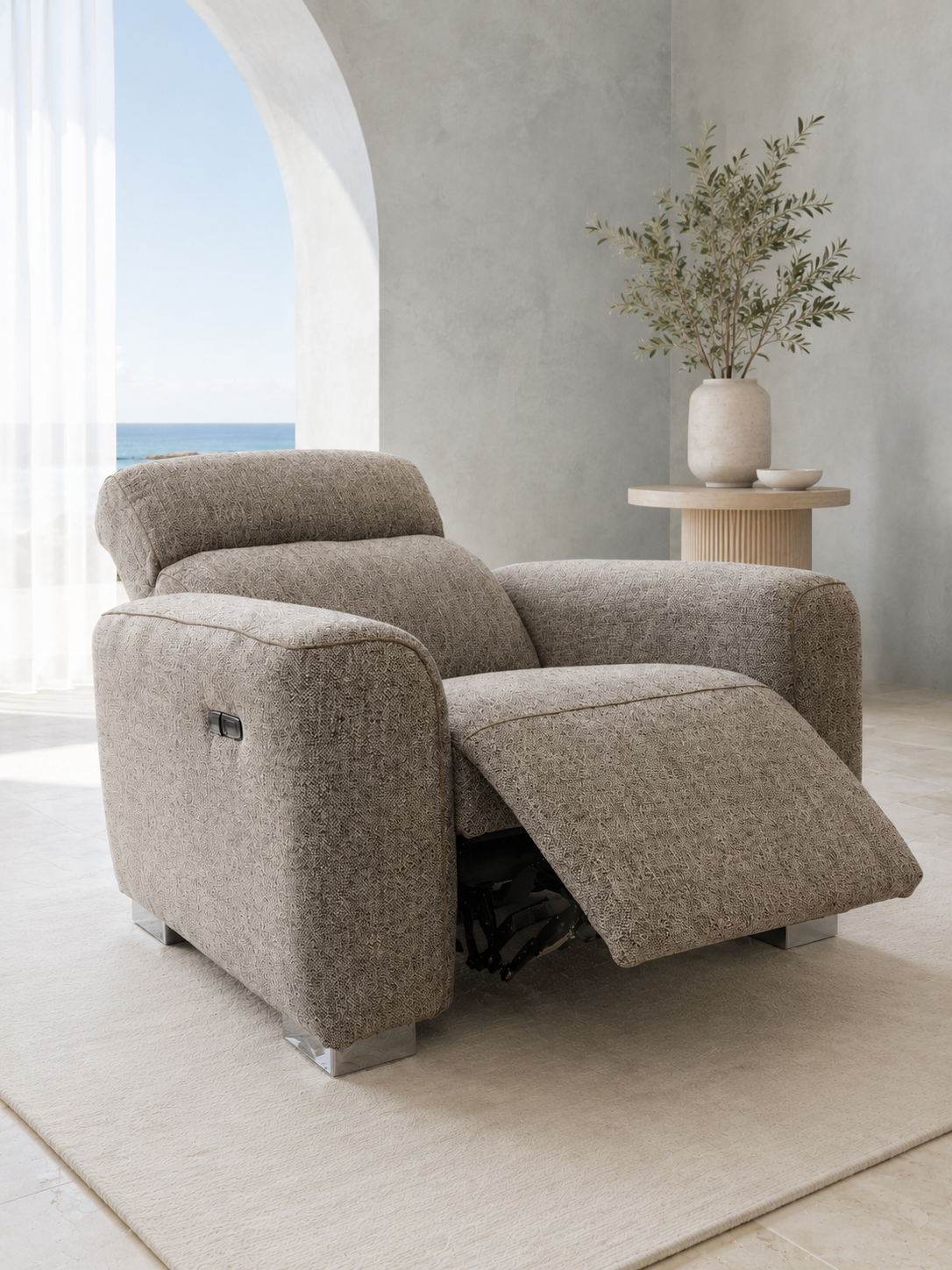 A modern upholstered recliner chair in a light taupe textured fabric, shown with the footrest extended in a bright, minimalist room with a side table and vase.