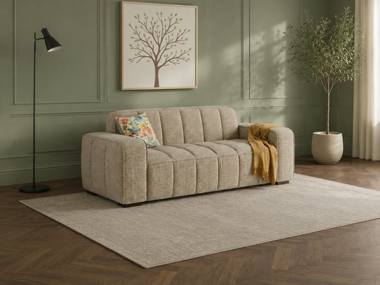 A modern tufted sofa in a light beige textured fabric, styled in a living room setting with a floral accent pillow, a mustard yellow throw blanket, a black floor lamp, a large area rug, and a potted tree against a sage green paneled wall.