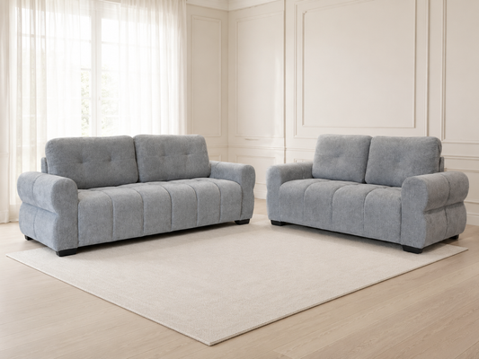 A light gray upholstered loveseat and a matching three-seat sofa placed on a large cream rug in a bright, elegant room with white walls, paneling, and sheer curtains.