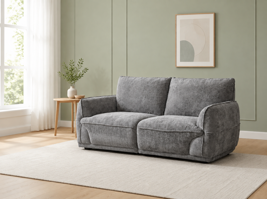A gray two-seater loveseat sofa in a textured fabric, styled in a modern living room with a light beige rug, a small wooden side table, and a sage green paneled wall.