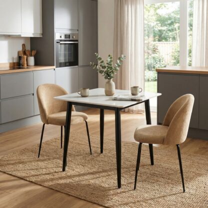 A modern dining set featuring a rectangular white table with black legs and two beige upholstered chairs, styled on a woven rug in a contemporary kitchen setting.