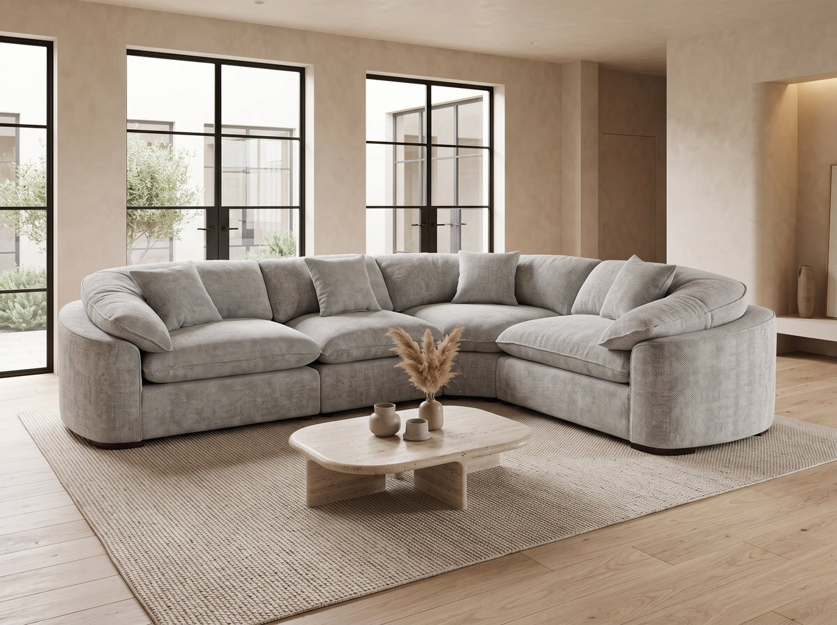 A light gray upholstered sectional sofa arranged in a U-shape in a modern living room with beige walls, large black-framed windows, a woven rug, and a small wooden coffee table.