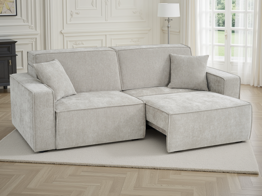 A light beige upholstered sectional sofa with a chaise lounge on the right side, shown in a bright, classic-style room with white paneled walls, a window with sheer curtains, and a light-colored rug.