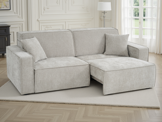 A light beige upholstered sectional sofa with a chaise lounge on the right side, shown in a bright, classic-style room with white paneled walls, a window with sheer curtains, and a light-colored rug.