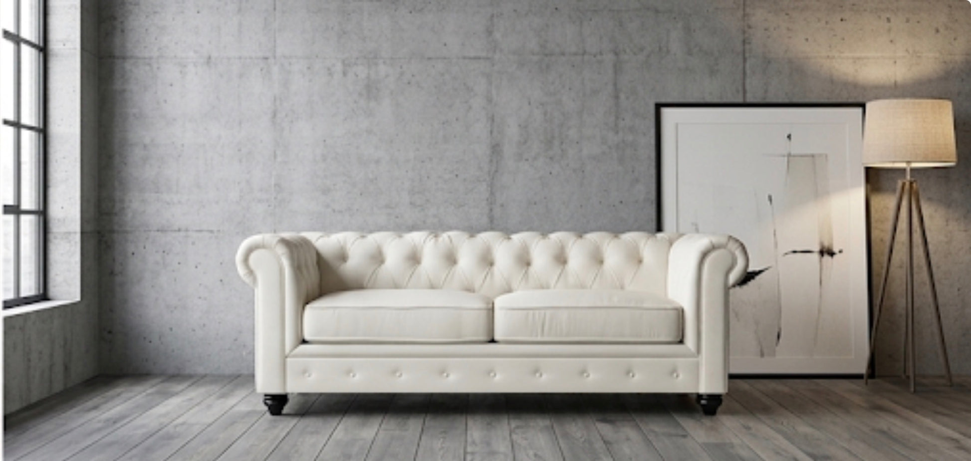 A cream-colored tufted Chesterfield sofa in a modern room with a concrete wall, wood floor, a window, a framed print, and a floor lamp.