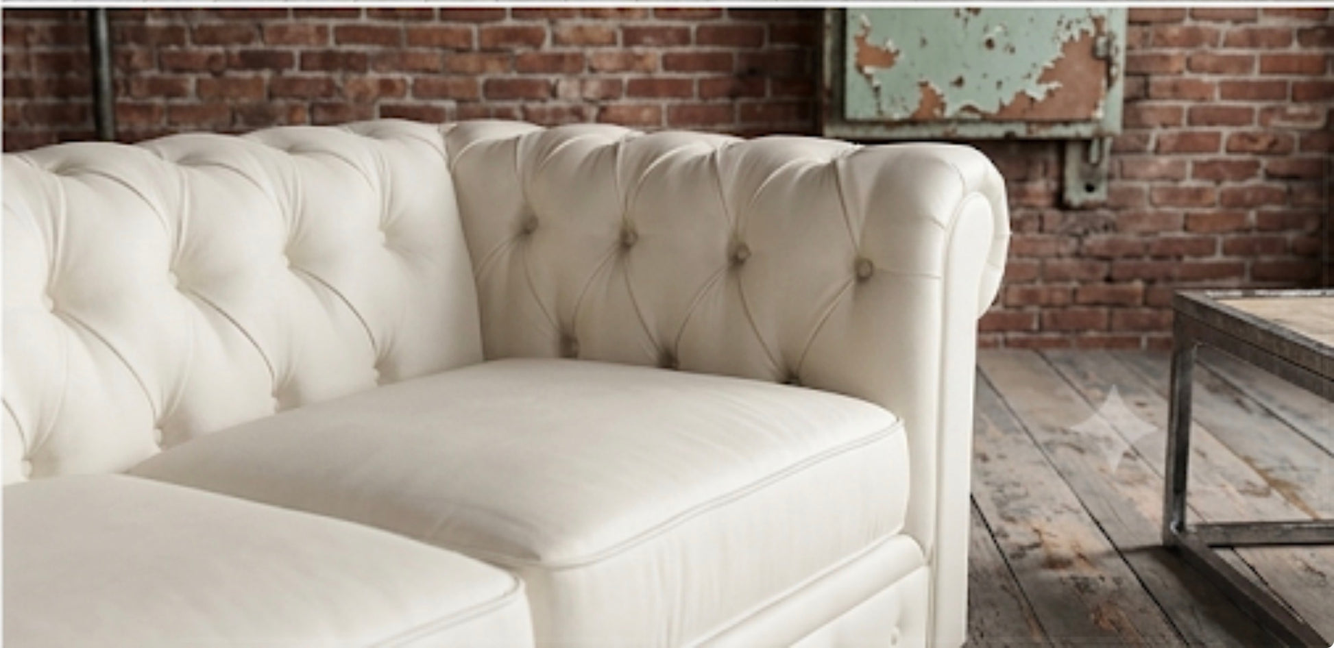 A cream-colored tufted leather sofa in a rustic room with a brick wall and wooden floor.
