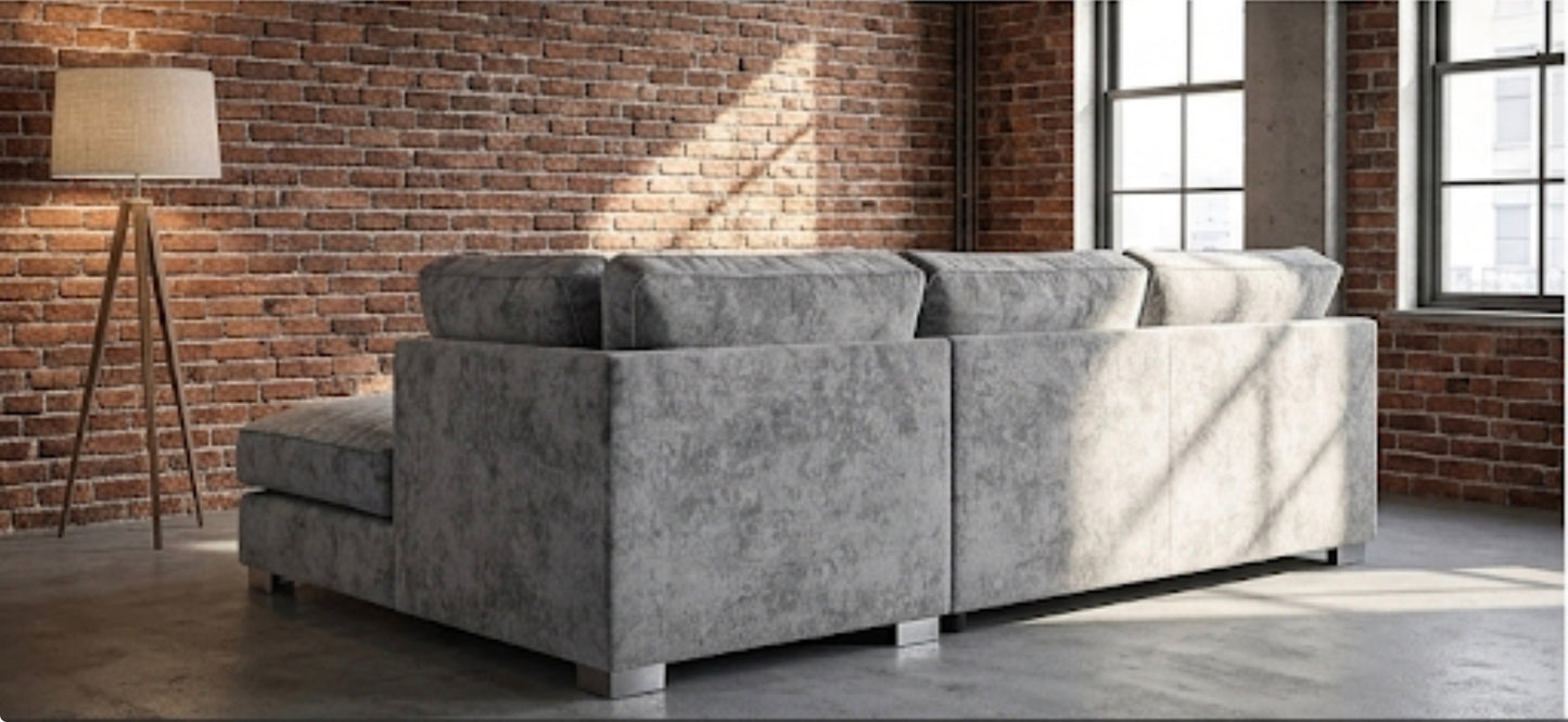 A light gray textured fabric sectional sofa with a left-facing chaise lounge, positioned in a modern loft room with a brick wall, floor lamp, and large windows.
