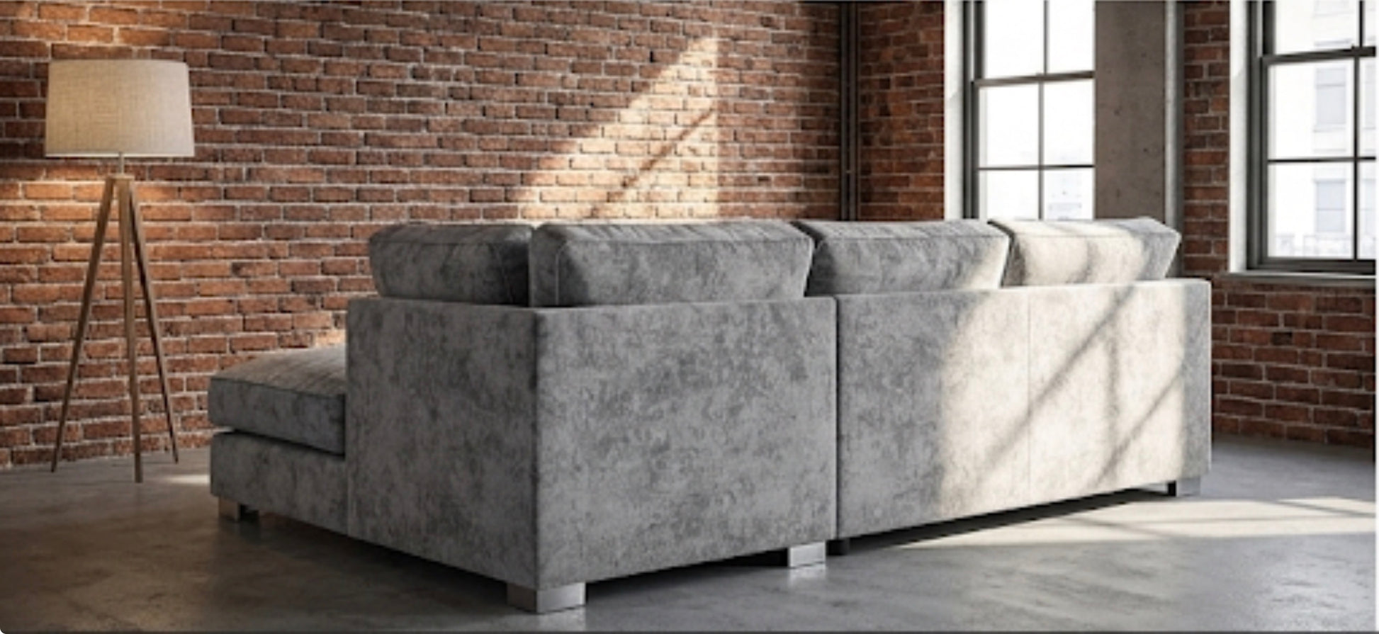 A light gray textured fabric sectional sofa with a left-facing chaise lounge, positioned in a modern loft room with a brick wall, floor lamp, and large windows.