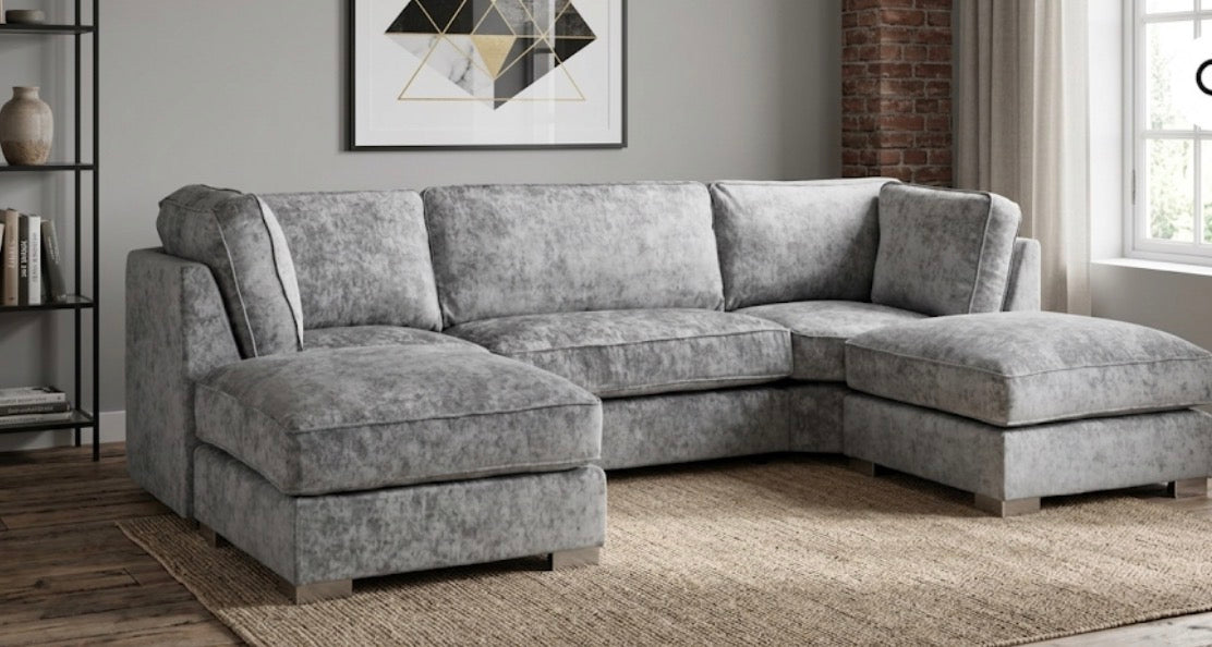 A light gray textured fabric sectional sofa with a left-facing chaise and a right-facing chaise, shown in a modern living room setting with a woven rug, brick wall, and window.