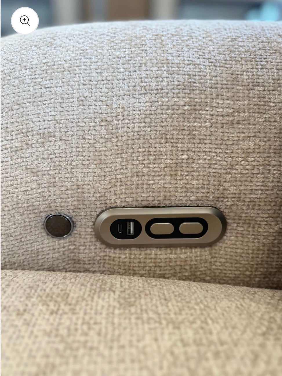 Close-up of a beige woven fabric surface with a small oval metallic button and a larger horizontal metallic control panel featuring a USB-C port and two touch buttons.
