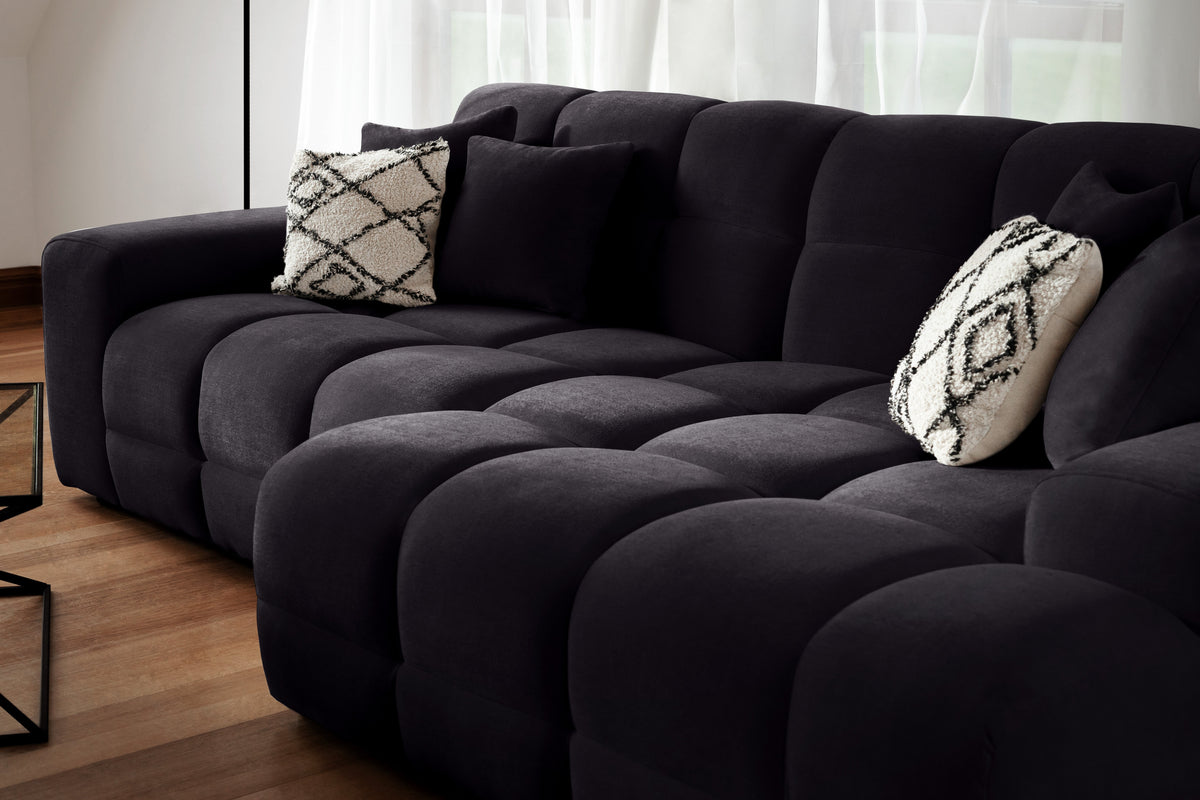Black sectional sofa with patterned cushions in a room with light-colored curtains.