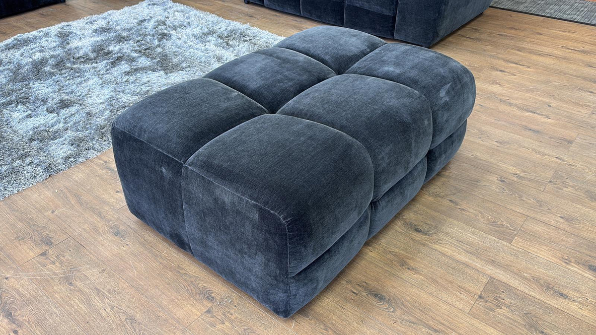 Black upholstered ottoman on a wooden floor with a rug in the background