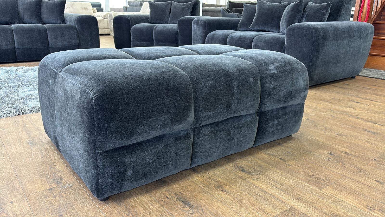 Black upholstered ottoman on a wooden floor with a sectional sofa in the background.
