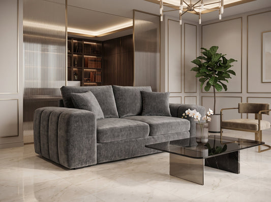 A modern gray velvet sofa with pleated armrests in a luxurious, contemporary living room featuring gold-trimmed walls, a glass coffee table, and a potted plant.