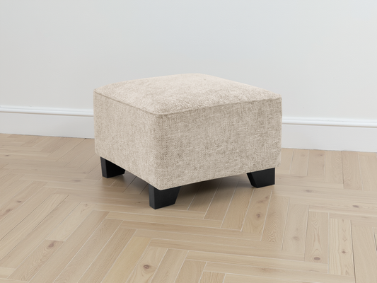 Beige ottoman with black legs on a wooden floor against a white wall