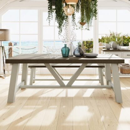 A rustic farmhouse dining table with a dark wood top and light gray painted legs, set in a bright, sunlit coastal dining room with an ocean view.