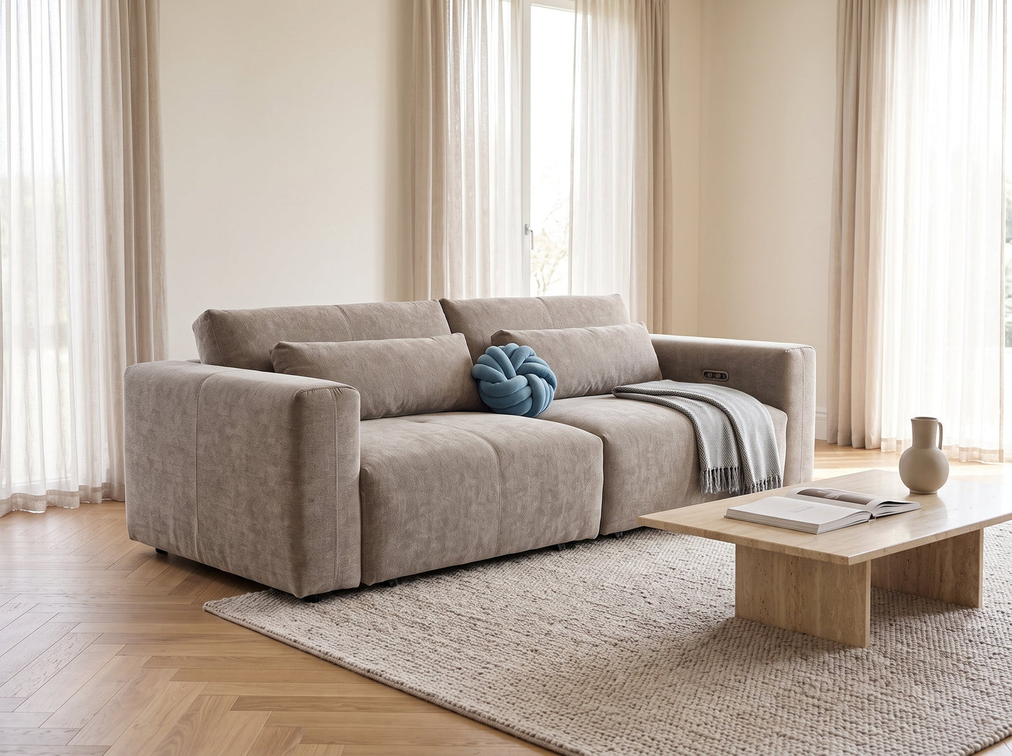 A modern beige fabric sofa with matching cushions in a bright, minimalist living room. A small blue knotted pillow and a gray throw blanket rest on the sofa, which is positioned on a textured beige rug next to a light wood coffee table.