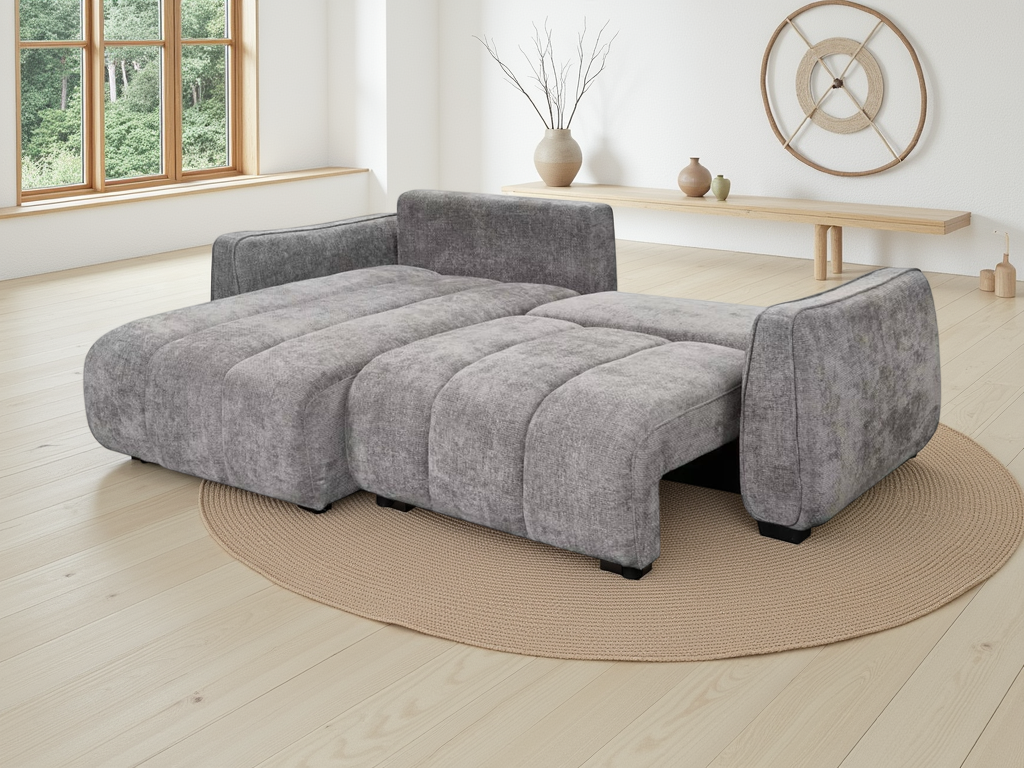 A modern grey fabric chaise lounge sofa with a padded footrest extension, shown in a bright, minimalist living room.