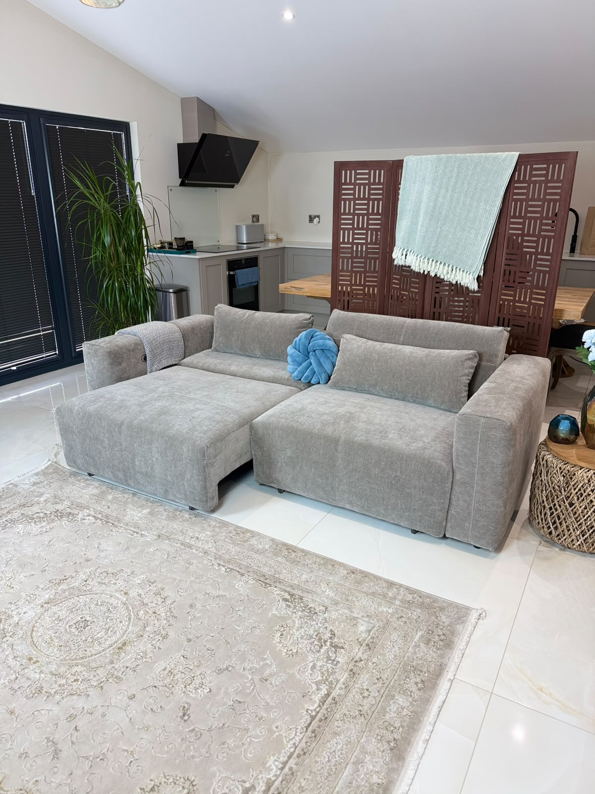 A light gray modular sectional sofa with a chaise lounge, styled in a modern living room with a beige patterned rug, a blue decorative pillow, and a kitchen area in the background.